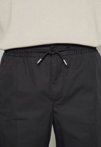 Pantalon noir léger avec une taille élastique et un cordon de serrage. Texture lisse avec des embouts métalliques aux extrémités du cordon.