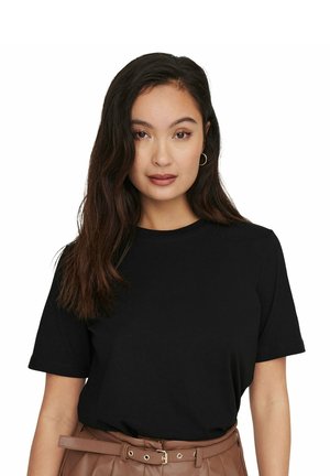 Giovane donna con lunghi capelli scuri, indossa una maglietta nera a maniche corte e una cintura marrone, posa su uno sfondo bianco uniforme.