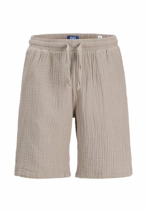 Chłopiec z kręconymi włosami, ubrany w beżową, krótką koszulę z wzorem i guziczkami, jasnobrązowe szorty, białe skarpetki i beżowe buty, stojący i uśmiechający się.