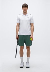 Hvit poloskjorte med en logo, grønne shorts og hvite sportssko. Holder en tennisracket i den ene hånden og en tennisball i den andre.