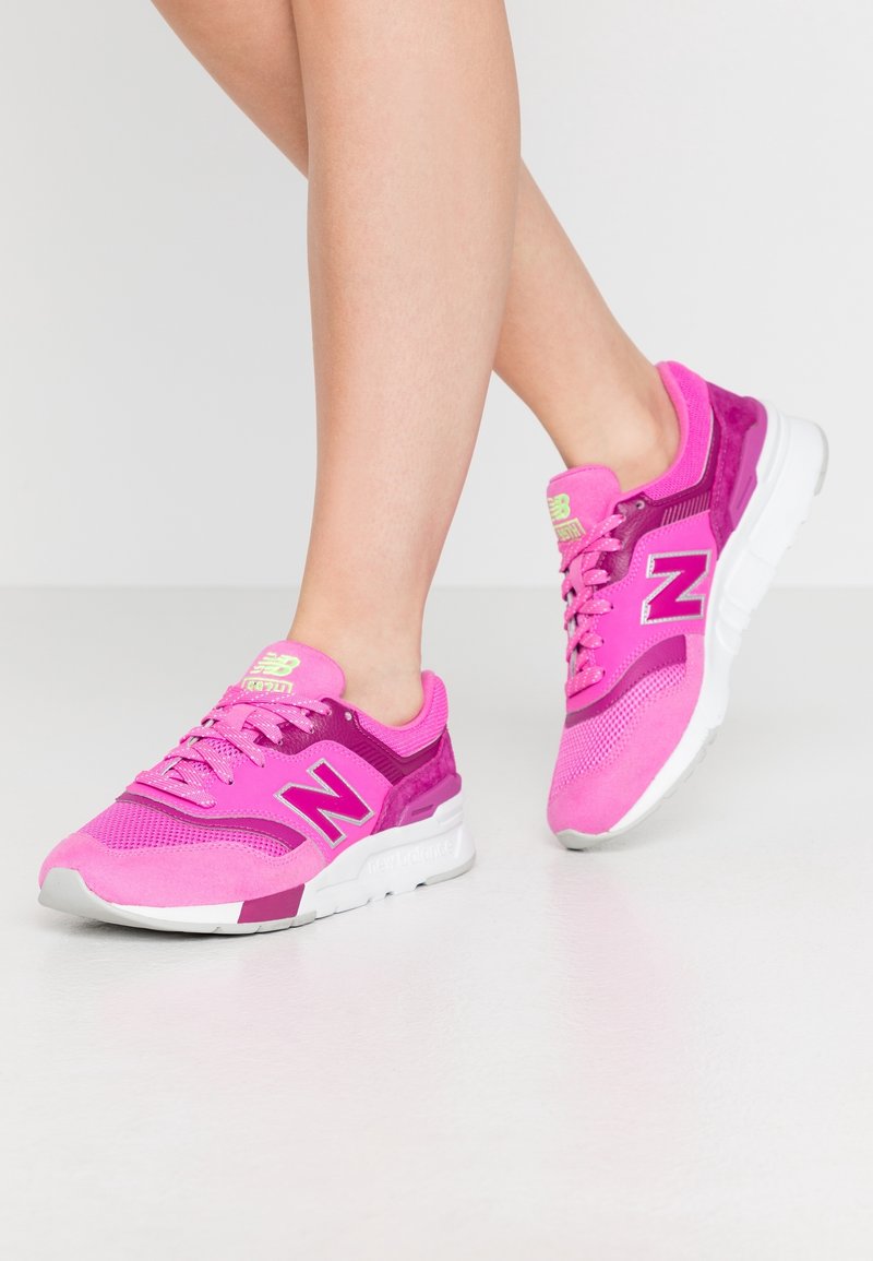Des jambes portant des baskets New Balance rose vif et violet avec des semelles blanches, posées sur un sol lisse gris clair.