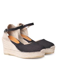 Melni kanvas wedge espadrillas ar potīšu siksniņām un austu jutu platformas zole, parādīti blakus viens otram uz balta fona.