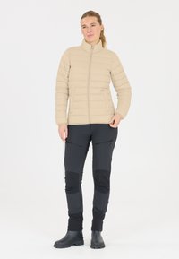 Beige isoleret jakke med lodrette sømme, høj krave og lynlås, parret med sorte bukser med lynlås lommer og knælapper.