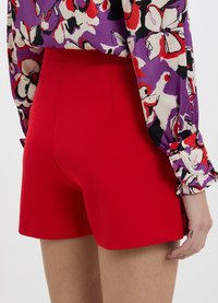 Shorts rossi a vita alta con una texture liscia, caratterizzati da linee pulite e una vestibilità sartoriale. Camicetta a fantasia floreale visibile sullo sfondo.