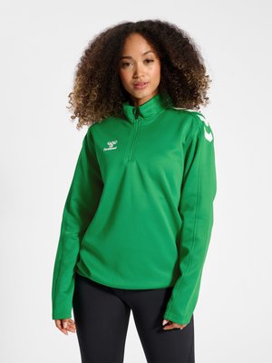Vrouw met krullend haar die een groene sporttrui met een kwart ritssluiting en zwarte legging draagt tegen een effen witte achtergrond.
