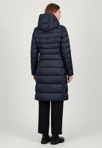 Cappotto imbottito blu navy con cappuccio, design a righe verticali, maniche lunghe e lunghezza fino a metà coscia. Abbinato a pantaloni neri e stivali.