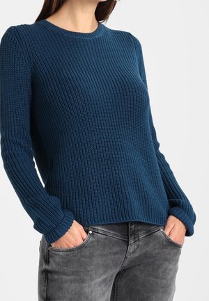 Femme porte un pull bleu en tricot à manches longues avec col rond et un jean gris délavé, les mains dans les poches avant.