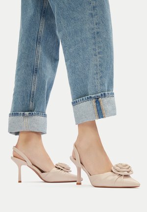 Beżowe szpilki na pasku z różanym akcentem, spiczastym noskiem i zakrzywionym obcasem typu szpilka, zestawione z podwiniętymi niebieskimi dżinsami.