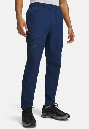 Man draagt marineblauwe cargobroek met elastische tailleband en zwarte sportschoenen, staand tegen een eenvoudige achtergrond.