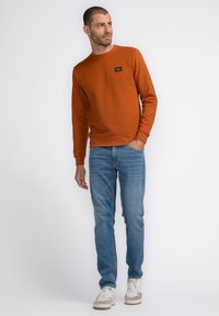 Bruine getextureerde sweatshirt met een ronde hals en logopatch, gecombineerd met lichtblauwe jeans en beige sneakers.