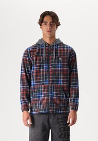 Zippered flannel jacket with red, blue, and black plaid pattern, gray hood, long sleeves, and logo patch on chest. Soft texture.