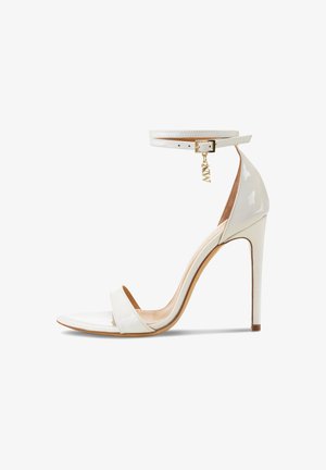 White patent leather heeled sandal with a thin ankle strap, gold buckle, and a charm. Features an open toe and a sleek, stiletto heel.