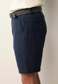 Tengeréskék vászonsort, slim fit fazonnal, oldalsó zsebekkel és egy hátsó zsebbel, fekete-fehér szőtt övvel párosítva.