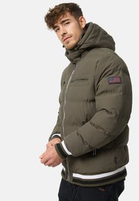 Olive green puffer jacket with a high collar, two zippered chest pockets, and striped ribbed cuffs and hem. Features an American flag patch.