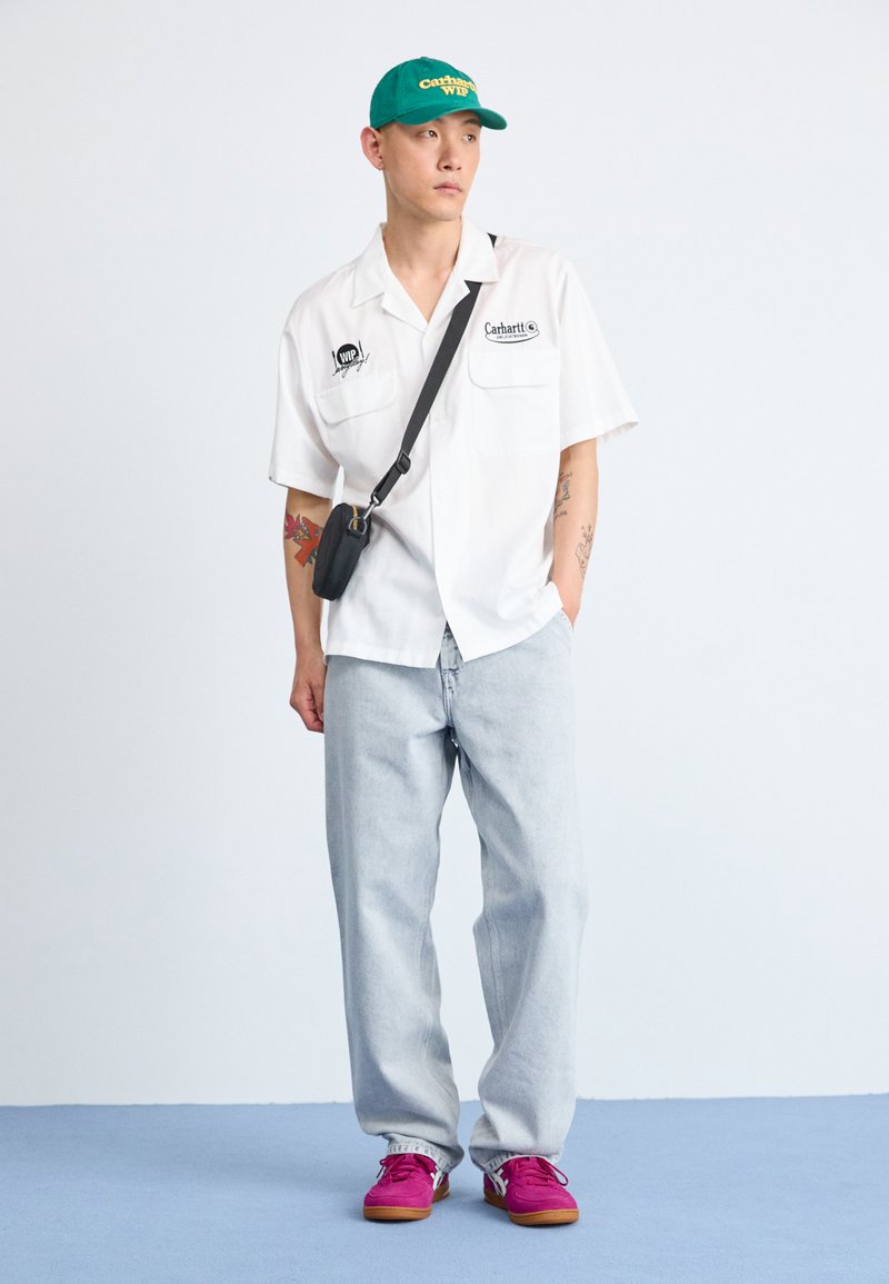 Chemise blanche à manches courtes, jeans bleu clair, casquette verte et baskets roses. Petit sac à épaule noir. Design simple et décontracté.