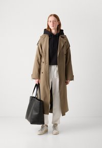 Lungo trench beige sopra una felpa nera, abbinato a pantaloni grigio chiaro e sneakers beige. Tiene una grande borsa a tracolla in pelle nera.