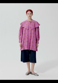 Femme portant un chemisier oversize à fleurs roses avec un grand col, un pantalon large noir coupé au-dessus de la cheville, un bandeau rouge et des ballerines argentées, debout sur fond blanc.