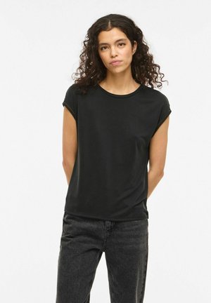 Göndör, sötét hajú nő fekete rövid ujjú felsőben és sötétszürke farmerral, kezét a háta mögé téve, egyszínű háttér előtt áll.