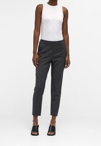 Pantalon gris ajusté avec une coupe slim, doté de poches latérales, de passants de ceinture et d'une texture lisse, associé à un top débardeur blanc ajusté et des sandales noires à enfiler.