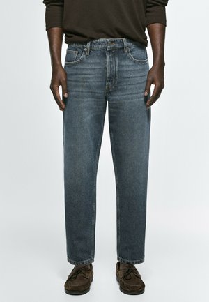 Mann trägt ausgewaschene blaue Jeans mit geradem Bein, dunkles Langarmshirt und braune Wildleder-Schnürschuhe, steht vor einem einfarbigen Hintergrund.
