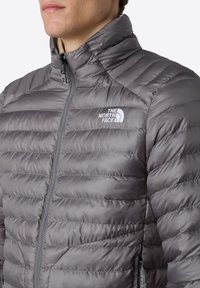 Šedá péřová bunda s vysokým límcem, prošívaným vzorem, plným zipem a logem The North Face na levé straně hrudníku. Měkká, hladká textura.