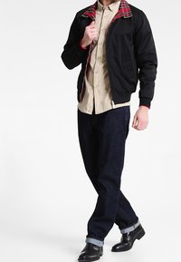 Giacca bomber nera con colletto a scacchi rossi, camicia beige sotto, jeans in denim scuro e stivali neri alla caviglia.