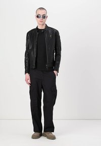 Giacca di pelle nera con cerniere, t-shirt nera, pantaloni cargo neri e scarpe beige. Gli accessori includono occhiali da sole e orecchini.