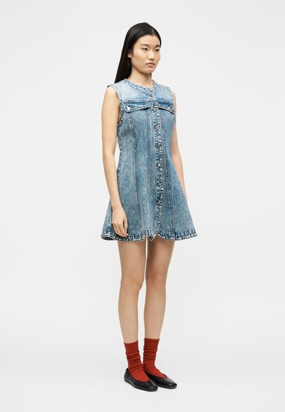 Νεαρή γυναίκα που φοράει μπλε τζιν φόρεμα χωρίς μανίκια με λεπτομέρειες με τρουκς, κόκκινες κάλτσες και μαύρα φλατ παπούτσια, στέκεται μπροστά σε λευκό φόντο.