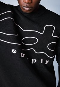 Czarny sweter dziany z białymi liniowymi grafikami oraz haftowanym na biało słowem "supply" na dole. Zbliżenie na teksturę materiału.