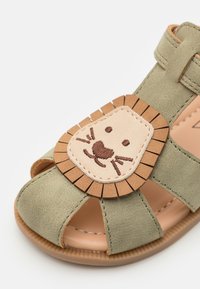 Grønn semsket sandal med åpen tå-design. Har en lysebrun løveansiktsapplikasjon med broderte detaljer og frynseakenter. Gummisåle.