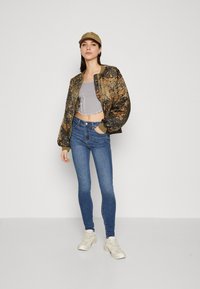 Brun og svart mønstret bomberjakke over en grå cropped topp, kombinert med blå skinny jeans og lyse joggesko, stående mot en ensfarget bakgrunn.