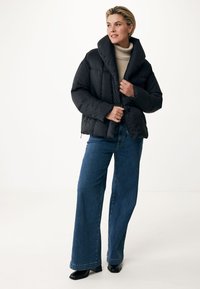 Doudoune noire avec un large col, présentant une texture matelassée et une fermeture éclair devant. Associée à un jean bleu évasé et des bottines à talons noires.
