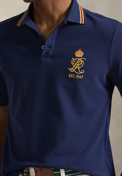 Μπλε σκούρο πόλο πουκάμισο από υφασμάτινη υφή, με γιακά με ρίγες σε κόκκινο και κίτρινο, κεντημένο λογότυπο με κορώνα και το λεπτομέρεια "EST. 1967".