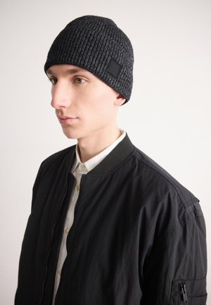 Jeune homme portant un bonnet en laine gris foncé, une veste bomber noire et une chemise blanche boutonnée, regardant légèrement vers la gauche.