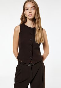 Brauner ärmelloser Vest mit hohem Kragen, ausgestattet mit sechs vorderen Knöpfen und einem schrägen Saum, kombiniert mit dunklen Hosen. Glatte Stofftextur.