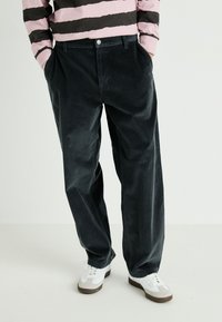 Pantalons en velours côtelé à jambes larges de couleur gris foncé avec des côtes verticales. Comprend des poches avant et une fermeture à bouton. Associés à des baskets blanches.