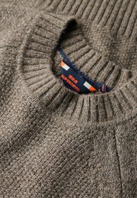 Pull en maille marron avec un col côtelé. Présente un tissu texturé et une étiquette affichant "SUPERDRY" en bleu marine et orange.
