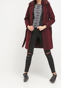 Bordeaux lang frakke med falsk pelskrave, parret med en sort mønstret top, slidte sorte skinny jeans og sorte ankelstøvler.