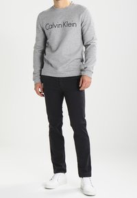 Sudadera gris con logo negro de "Calvin Klein", combinada con pantalones negros y zapatillas blancas. Diseño simple, tejido ligero y corte relajado.