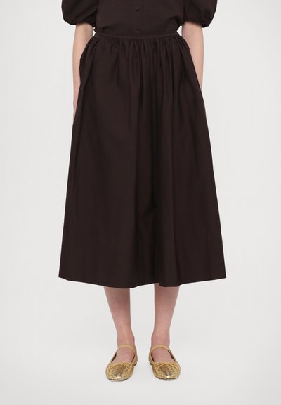 Brown, cotton, mid-length skirt with an elastic waistband and side pockets, paired with braided gold flat shoes featuring yellow accents.