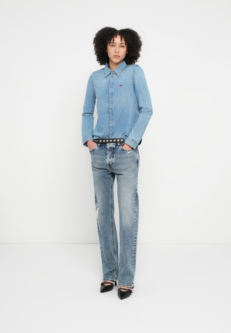 Lyseblå denimskjorte med knaplukning, lange ærmer og rød logo. Kombineret med slidte lyseblå jeans og sorte spidse sko.