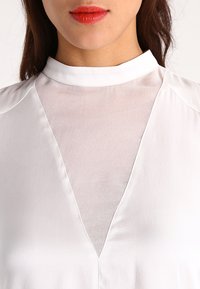 Weiße Bluse mit einem transparenten oberen Paneel im V-Ausschnitt, glatter Textur und hochgeschlossenem Kragen. Keine sichtbaren Muster oder Beschläge.