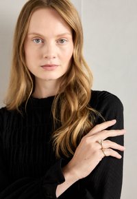 Goldring mit einem klaren Stein, strukturiertes Gewebe eines schwarzen Langarmoberteils, Model positioniert vor einem hellgrauen Hintergrund.