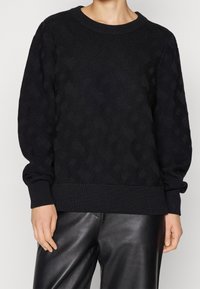 Suéter negro de punto con patrón texturizado, cuello y puños acanalados, corte holgado, combinado con pantalones de cuero negro de cintura alta.