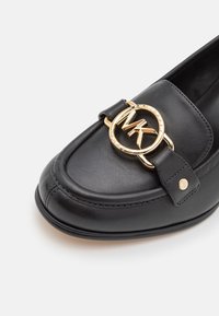 Mocassin en cuir noir avec un logo circulaire doré proéminent portant les initiales "MK", orné d'une sangle et de détails en métal doré. Texture lisse.