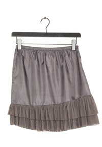 Šedá vícevrstvá sukně vyrobená z lehkého materiálu, s volánkovým lemem a elastickým pasem, zavěšená na černém dřevěném ramínku.