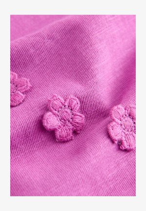 Svetlo rožnata tkanina z mehkimi teksturami, na kateri so trije vezeni cvetovi v temnejši rožnati barvi. Dizajn prikazuje gladek in enakomeren vzorec pletenine.