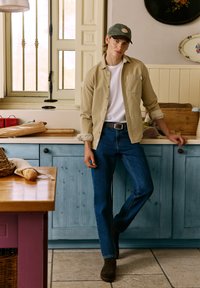 Beige button-up shirt over a white t-shirt, paired with blue jeans and brown boots. Standing in a kitchen with blue cabinetry.
