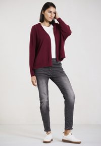 Burgunderfarbener Strickcardigan über einem weißen Oberteil, kombiniert mit grauen Slim-Fit-Jeans und weißen Sneakers mit braunen Akzenten. Zeitlose Textur, lockere Passform.
