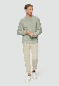 Mann in einem hellgrauen Kabelstrickpullover, beige hochgekrempelten Hosen und weißen Sneakers, der vor einem schlichten hellen Hintergrund steht.